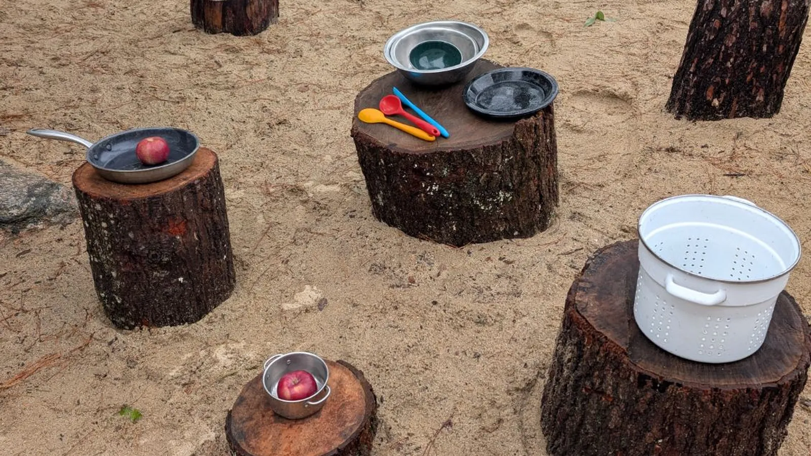 A mud kitchen of tree stumps with pans, bowls, spoons, and apples set up in the sand for TimberNook at Blossom Station